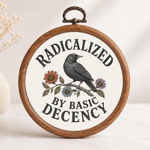 Op de afbeelding: Een ronde houten borduurring met witte stof waarop een illustratie van een zwarte kraai op een bloemrijke tak staat. De tekst RADICALIZED BY BASIC DECENCY is in zwarte hoofdletters rondom de centrale vogelafbeelding gedrukt.
