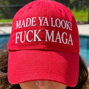 May include: A red baseball cap with the text "MADE YA LOOK! FUCK MAGA" in white. The cap is worn low, partially obscuring the wearer's face. The background includes a swimming pool and greenery.