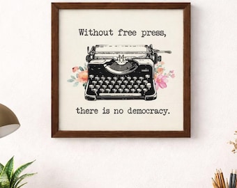 May include: A framed print with a vintage typewriter illustration and floral details. The text reads, "Without free press, there is no democracy." The artwork is displayed on a wooden desk with a lamp, plant, and other office supplies.