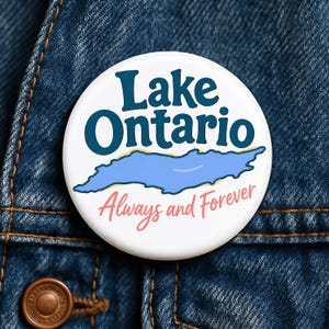 May include: A round white pinback button featuring the dark blue text Lake Ontario above a light blue illustration of the lake. Below the lake graphic, the text Always and Forever is written in a cursive pink font on a white background.