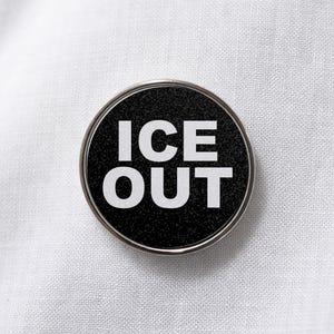 May include: A circular pin-back button featuring a black, textured, glittery background with the words ICE OUT printed in bold, white, sans-serif capital letters. The button has a thin, polished silver-toned metal rim around the edge.