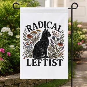 May include: A white rectangular garden flag featuring a black cat illustration centered among colorful wildflowers. The text RADICAL is printed in black above the cat, and the text LEFTIST is printed in black below the cat on the white fabric.