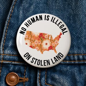 Puede incluir: Botón blanco con el texto "NO HUMAN IS ILLEGAL ON STOLEN LAND". El botón presenta un diseño floral del mapa de Estados Unidos en naranja, rojo y blanco. El botón está sobre un fondo de mezclilla azul.