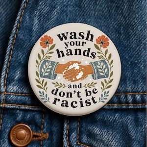 Könnte beinhalten: Ein runder Ansteckbutton mit cremefarbenem Hintergrund und einer Illustration von zwei Händen, die sich mit Seife waschen. Der Text lautet wash your hands and don't be racist. Das Design zeigt orangefarbene Blumen und grüne Blätter an den Seiten.