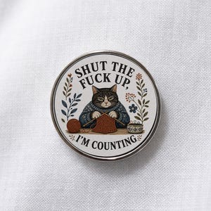 May include: A circular pin featuring an illustration of a cat wearing a blue sweater and knitting with brown yarn. The text reads SHUT THE FUCK UP, I'M COUNTING. The design includes floral elements, a ball of yarn, and a small bowl of knitting needles.