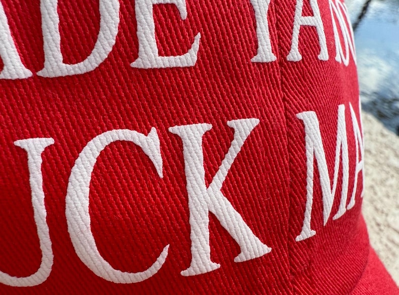 May include: Red baseball cap with white embroidered text. The hat's fabric has a textured appearance. The text on the hat reads "JUCK MA".