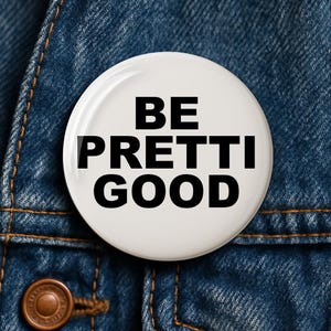 May include: A white button with the words "BE PRETTI GOOD" in bold black letters. The button is set against a blue denim background, with visible stitching and a button.