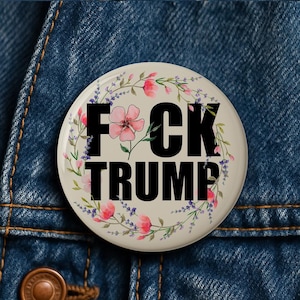 May include: A round, cream-colored button with the text "F*CK TRUMP" in bold black letters. The text is surrounded by a floral wreath of pink and purple flowers and green leaves. The button is set against a blue denim background.
