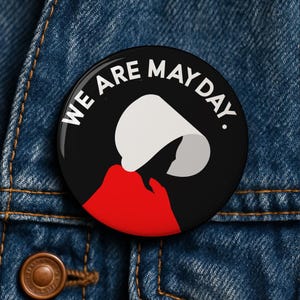 May include: A round black button with the text "WE ARE MAYDAY." in white. The button features a graphic of a white bonnet and red cloak on a black background, set against a denim jacket.