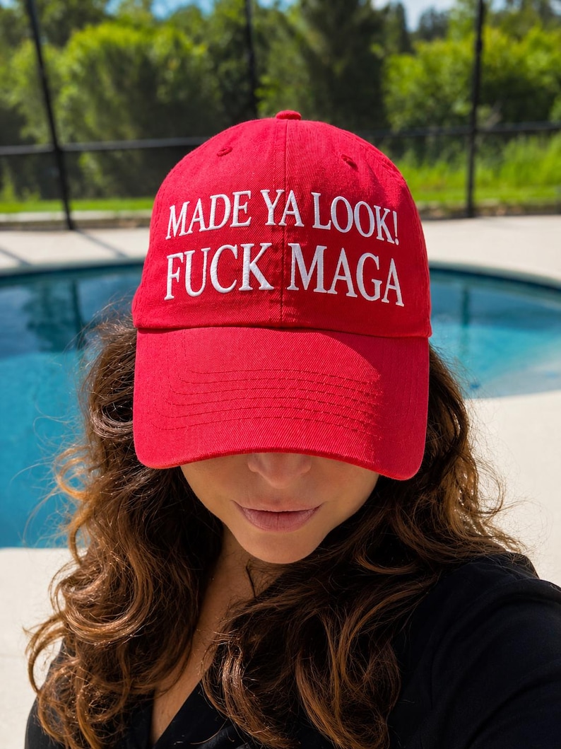 May include: A red baseball cap with the text "MADE YA LOOK! FUCK MAGA" in white. The cap is worn low, partially obscuring the wearer's face. The background includes a swimming pool and greenery.