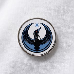Può includere: Una spilla metallica circolare raffigurante un colimbo nero con occhi rossi e ali spiegate, centrato su una forma a mezzaluna azzurra. Sopra l'uccello si trova una piccola stella azzurra a quattro punte su sfondo bianco.
