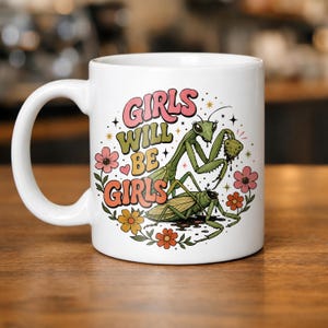 May include: A white ceramic mug featuring a colorful graphic of two green praying mantises surrounded by pink, orange, and yellow flowers. The text "GIRLS WILL BE GIRLS" is printed in a retro, wavy font above the insects, with small star accents.