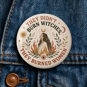 May include: A round, white button with the text "THEY DIDN'T BURN WITCHES, THEY BURNED WOMEN." The design features a woman's silhouette, flames, sun, moon, and floral elements in brown, beige, and orange tones. The button is set against a blue denim background.