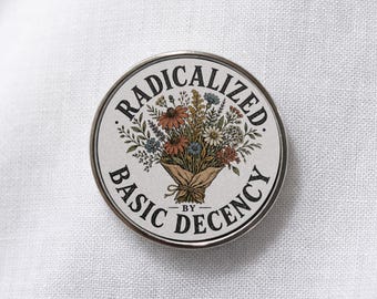 May include: A circular pin-back button featuring a central illustration of a wildflower bouquet wrapped in brown paper. The text RADICALIZED BY BASIC DECENCY is printed in black capital letters in a circular arc around the floral design.
