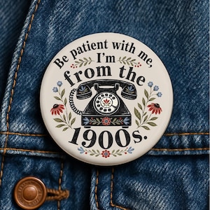 May include: A round pinback button with a cream background featuring a black rotary telephone illustration, floral accents, and the text: Be patient with me, I'm from the 1900s. The design includes red and blue flowers and green leaves surrounding the phone.