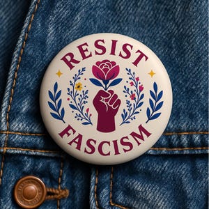 May include: A round, white button with the words "RESIST FASCISM" in burgundy lettering. The button features a burgundy fist holding a flower, surrounded by floral and leaf designs in blue, yellow, and pink. The button is set against a blue denim background.
