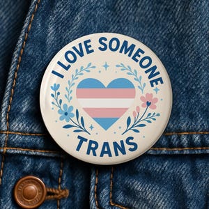 May include: A circular badge with the words "I LOVE SOMEONE TRANS" in blue. The design includes a heart in the transgender flag colours, framed by floral and leaf motifs, on a cream-coloured backdrop.