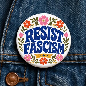 May include: A circular pinback button with a white background featuring the blue text RESIST FASCISM in a bold, wavy font. The design is surrounded by a colourful floral wreath with pink and orange flowers, green leaves, and a small yellow banner.