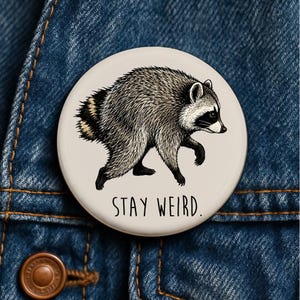 Op de afbeelding: Een ronde button met een gedetailleerde zwart-witillustratie van een lopende wasbeer. De achtergrond van de button is lichtcrème en de tekst "STAY WEIRD." is in een eenvoudig zwart schreefloos lettertype onder de wasbeer gedrukt.