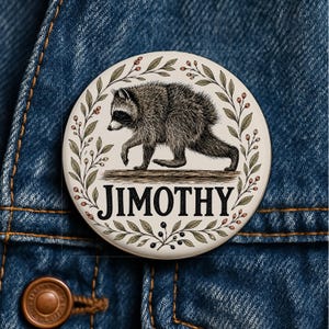 Op de afbeelding: Een ronde button met een crèmekleurige achtergrond en een gedetailleerde illustratie van een wasbeer die op een boomstam loopt. De wasbeer is omringd door een krans van groene bladeren en kleine rode bessen. De naam JIMOTHY staat in het zwart.