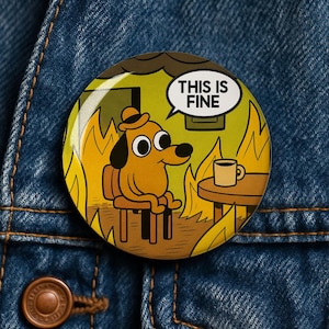 Könnte beinhalten: Ein runder Ansteckbutton mit einer Cartoon-Illustration eines orangefarbenen Hundes mit Hut, der an einem Tisch mit einer Kaffeetasse sitzt, während er von Flammen umgeben ist. Eine Sprechblase über dem Hund enthält den schwarzen Text, THIS IS FINE.