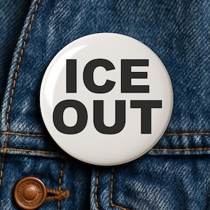 May include: A white button with the words "ICE OUT" in bold, black capital letters. The button is set against a blue denim background. The button is round and appears to be a pin or badge.