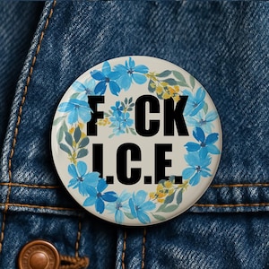 Puede incluir: Un botón redondo con el texto "F*CK I.C.E." en negro, rodeado por una corona de flores azules y detalles amarillos. El botón está sobre un fondo de mezclilla.
