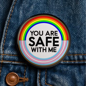 May include: A circular pin-back button featuring a white center with the black text YOU ARE SAFE WITH ME. The design is surrounded by a rainbow arc on the top half and a trans pride flag color arc on the bottom half, framed by a thin black border.