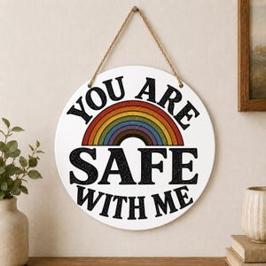 May include: A round white wall sign hanging from a jute rope. The sign features a colorful rainbow graphic in the center with the black text "YOU ARE" arched above the rainbow and "SAFE WITH ME" printed in large black letters below it.