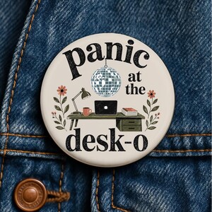 May include: A round pinback button with a cream background featuring the black text "panic at the desk-o". The design includes a silver disco ball, a desk with a laptop, a lamp, a mug, and books, flanked by two floral stems with orange flowers.