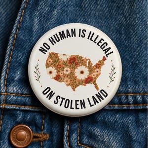 May include: A round pinback button with a white background featuring a map of the United States filled with a floral pattern in shades of orange, red, and white. The text "NO HUMAN IS ILLEGAL ON STOLEN LAND" is printed in black capital letters.