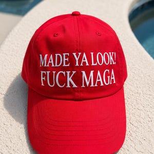 May include: A red baseball cap with the text "MADE YA LOOK! FUCK MAGA" in white block letters. The cap has a curved brim and a button on top. The background is a light-colored surface.