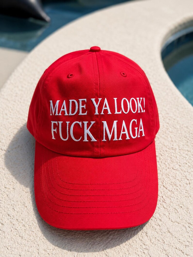 May include: A red baseball cap with the text "MADE YA LOOK! FUCK MAGA" in white block letters. The cap has a curved brim and a button on top. The background is a light-colored surface.