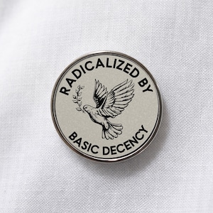 Op de afbeelding: Een ronde, zilverkleurige pin met de tekst "RADICALIZED BY BASIC DECENCY" rond een duif die een olijftak draagt. De duif is zwart en wit, tegen een lichtgrijze achtergrond.