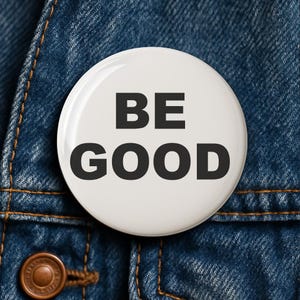 May include: A white button with the words "BE GOOD" in bold, black letters. The button is set against a blue denim background, with visible stitching and a metal button.