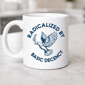 Könnte beinhalten: Weißer Keramikbecher mit einer blauen Illustration einer Taube, die einen Olivenzweig trägt. Der Becher trägt den Text "RADICALIZED BY BASIC DECENCY" in einem Kreis um die Taube.