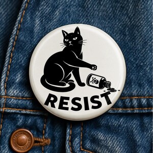 May include: A circular pinback button with a white background featuring a black illustration of a cat knocking over a small bottle labeled with a skull and crossbones symbol. The word RESIST is printed in bold black capital letters at the bottom.