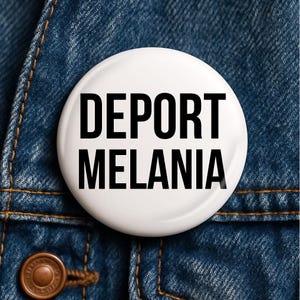 May include: A white badge with the words "DEPORT MELANIA" in bold black letters. The badge is set against a blue denim jacket, with the badge's edge slightly visible. The jacket's texture and stitching are clearly visible.
