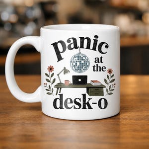 May include: A white ceramic mug featuring a graphic of a desk with a laptop, lamp, and mug, topped by a disco ball. The text reads "panic at the desk-o" in black lettering. Floral sprigs with orange flowers flank the desk on both sides of the mug.
