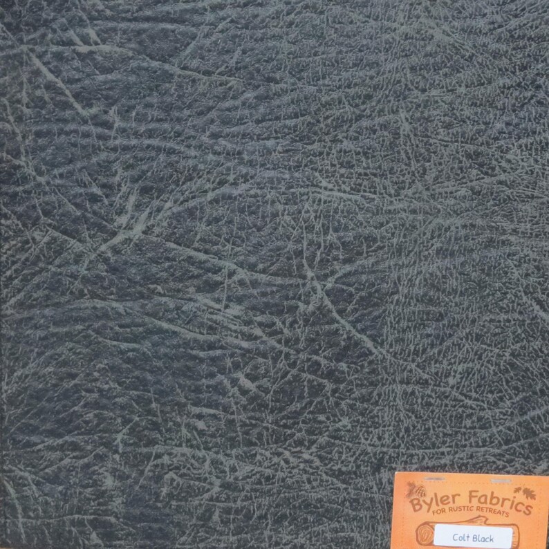 May include: Close-up of a dark grey textured fabric with a subtle green undertone. The fabric has a leather-like appearance and is labelled "Colt Black" by "Byler Fabrics for Rustic Retreats".