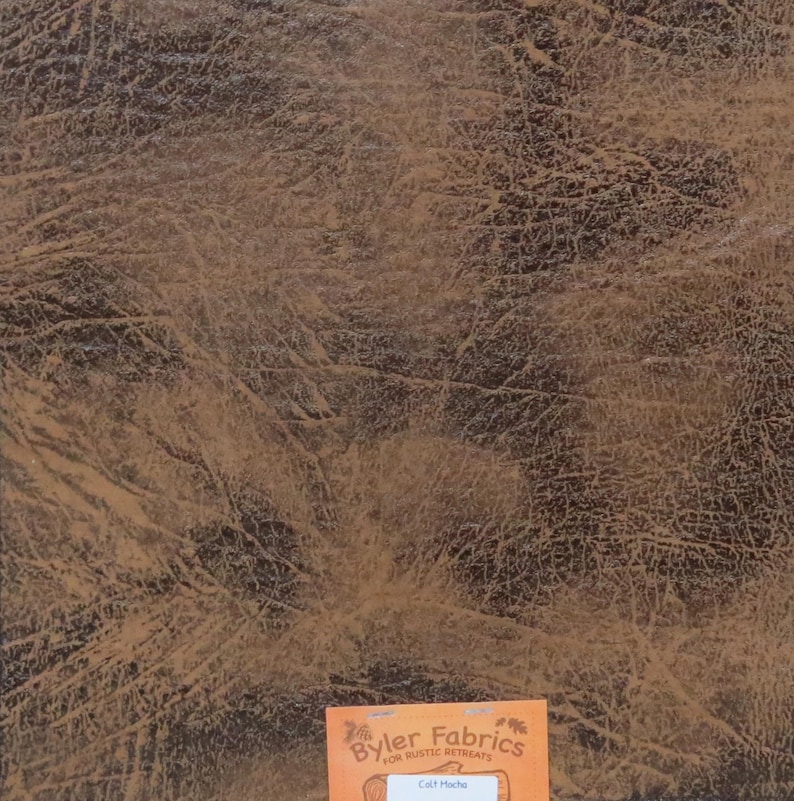 May include: Close-up of a brown faux leather fabric with a distressed, textured look. The fabric is labelled "Byler Fabrics for Rustic Retreats" and "Calf Mocha".