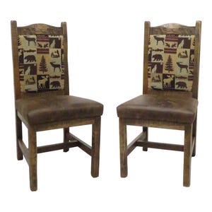 May include: Two wooden chairs with brown leather seats and backrests featuring a fabric panel with a repeating pattern of bears, moose, deer, fish, and pine trees in shades of brown, tan, and cream. The chairs have a sturdy frame and straight legs.