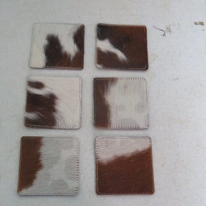 May include: Six brown and white cowhide coasters with stitched edges. The coasters are arranged in two rows of three.