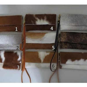 May include: A collection of nine leather clutch wallets with wrist straps. The wallets feature various cowhide patterns, including solid brown, white and brown, and gray. Each wallet has a rectangular shape with a fold-over flap.