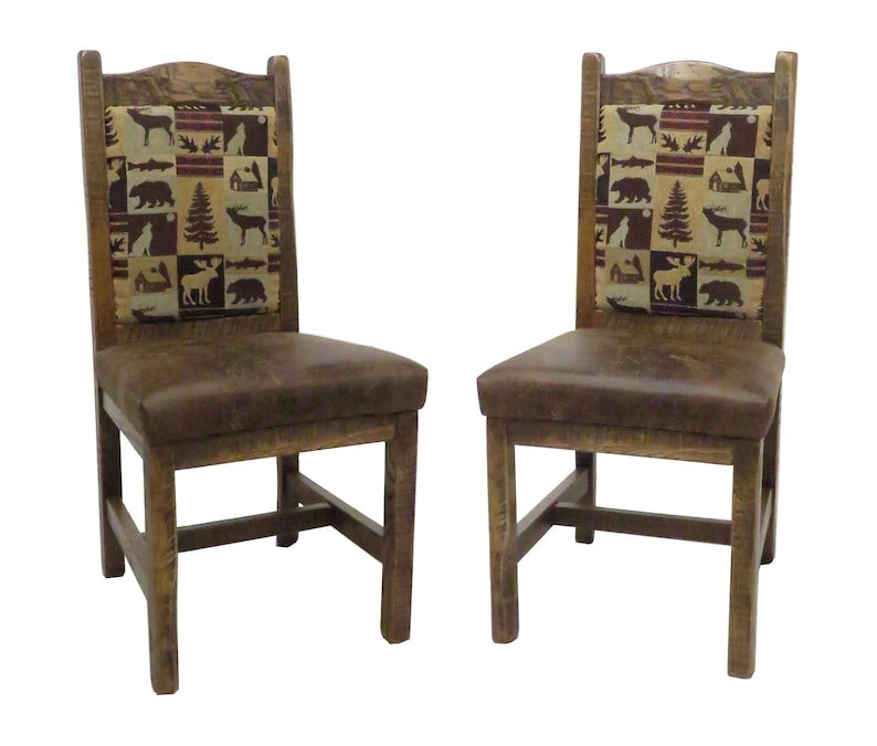 May include: A pair of wooden chairs with brown leather seats and high backrests. The backrests feature a rectangular fabric panel with a repeating pattern of bears, moose, deer, fish, and pine trees in shades of brown, tan, and dark red.