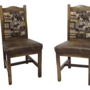 May include: Two wooden chairs with dark brown leather seats and high backrests. The backrests feature a rectangular fabric panel with a patchwork pattern depicting wildlife including deer, a bear, and a moose, set against a forest background.