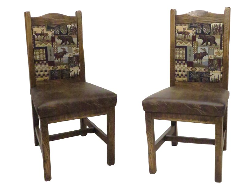 May include: Two wooden chairs with dark brown leather seats and high backrests. The backrests feature a rectangular fabric panel with a patchwork pattern depicting wildlife including deer, a bear, and a moose, set against a forest background.