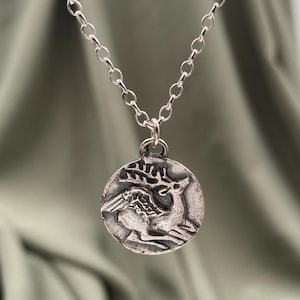 Puede incluir: Un collar de cadena de plata con un colgante redondo que presenta una imagen en relieve detallada de un ciervo con cuernos y alas.