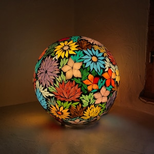May include: A round, mosaic glass lampshade with a floral design in shades of blue, yellow, red, green, and purple. The lampshade is illuminated from within, casting a warm glow.