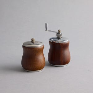 May include: A pair of kitchen tools featuring a wooden salt shaker and a wooden pepper mill. Both items have a curved, hourglass-shaped body made of dark brown wood, accented with polished silver-tone metal lids and a manual crank handle.